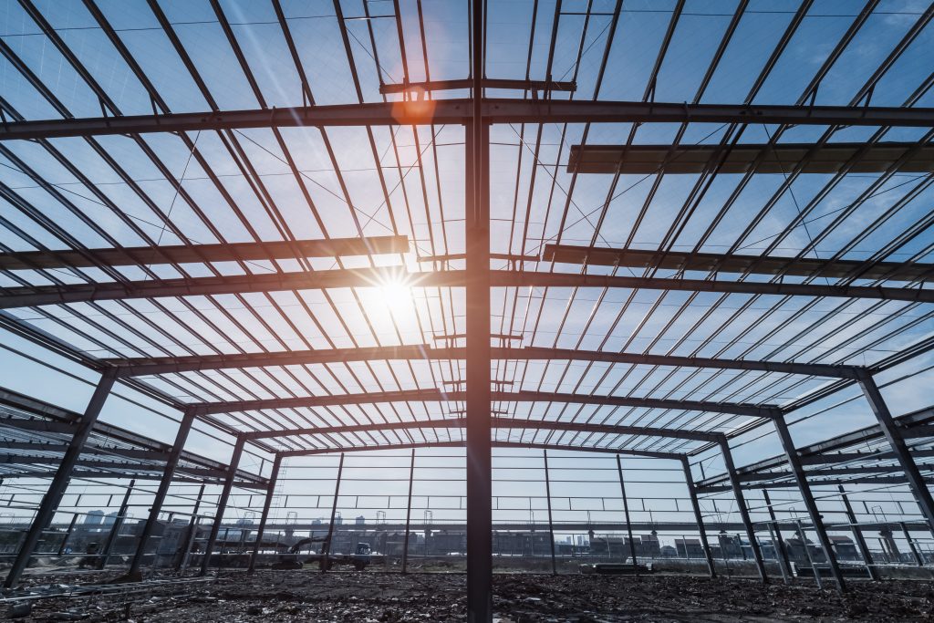 A low-angle perspective showcases the structural framework of a building under construction. Steel beams form a geometric pattern against the bright blue sky, with the sun shining through the open roof.